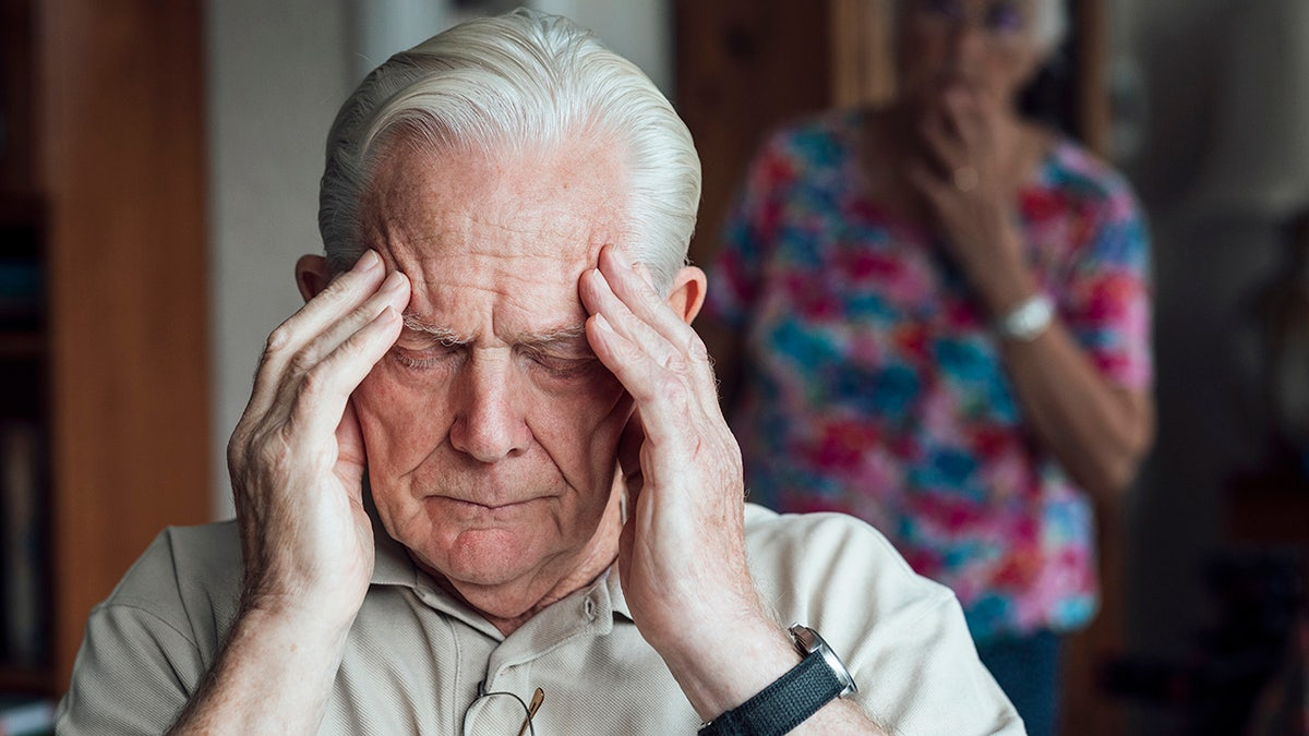 Senior man who has a chronic illness/Alzheimer's disease sitting in the living room in his north home -east of England. Is keeping the front with a negative expression, experiencing anxiety/pain. His wife is out of focus behind him, looking at him with concern.